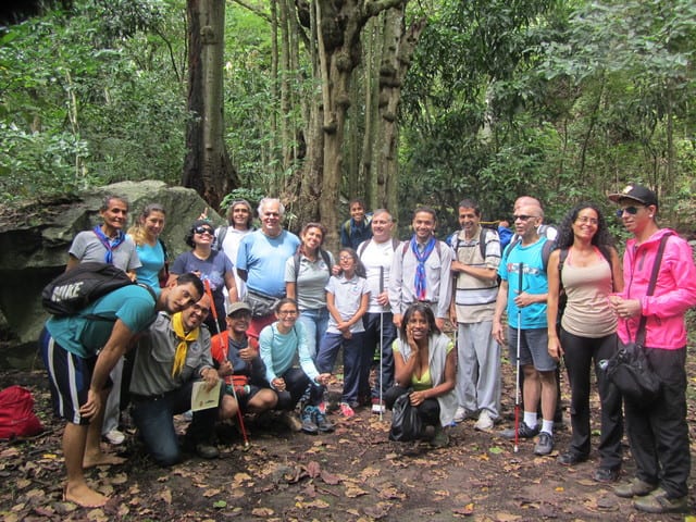 Grupo de PCD visual con sus guías (20 personas), todos muy sonrrientes, en el camino a Quebrada Quintero, con grandes árboles y rocas de fondo, durante la segunda caminata al Ávila.